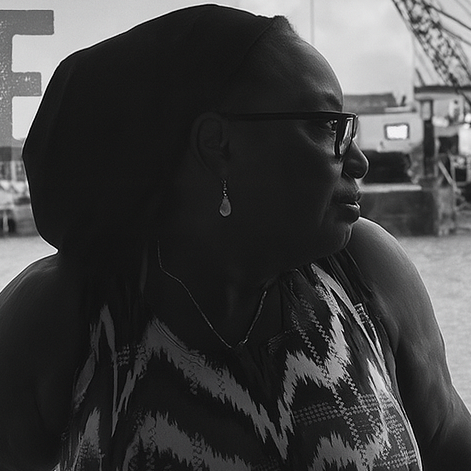 A Black woman wearing a headscarf and patterned sleeveless dress looks pensively to her right. She is seated in front of a window with blurred writing and light coming through. The image is in black and white, highlighting her profile and calm presence.